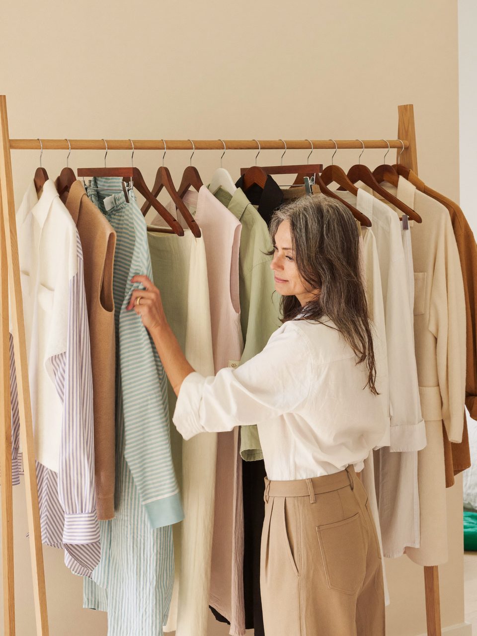 A woman in her forties choosing a shirt from a rail of neutral clothes in a bright bedroom